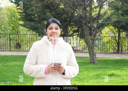 Frau lächelt im Freien, hält Telefon. Trägt eine gemütliche weiße Fleecejacke. Grünes Gras und Bäume hinter ihr. Metallzaun im Hintergrund sichtbar. Helles, nat Stockfoto