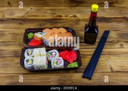 Sushi-Brötchen in Plastikkisten, Sojasauce und Essstäbchen auf Holztisch. Sushi zum Mitnehmen oder zur Lieferung von Sushi in Kunststoffbehältern Stockfoto