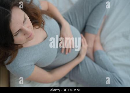Schwangere Frau auf dem Bett mit Händen auf dem Bauch aus einer Oberblicksperspektive. Erwartende Mutter schaut nach unten, umarmt die Mutterschaft und die glückliche Erwartung. zukünftige Mutter in einem ruhigen Schlafzimmer, die sich zufrieden und verbunden fühlt Stockfoto