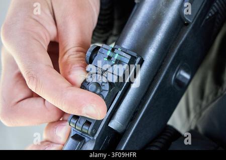 Nahaufnahme zeigt eine Hand, die das faseroptische Rückvisier am Gewehr einstellt. Der Finger dreht eine nummerierte Skala für eine präzise Zielkalibrierung. Outdoor-Schießausrüstung Stockfoto