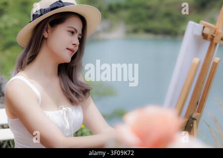 Eine anmutige junge Frau in einem Sommer-Outfit erschafft Kunstwerke auf einer Staffelei im Freien neben einem ruhigen See, perfekt für kreative Lifestyle-Themen. Stockfoto