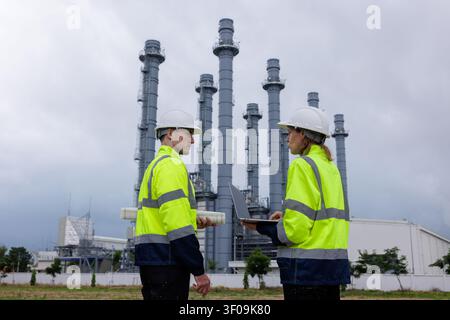 Zwei Ingenieure in Sicherheitshelmen und Warnkleidung arbeiten gemeinsam an Projektplänen mit einem Laptop und Bauplänen in einem Industriekraftwerk. Stockfoto