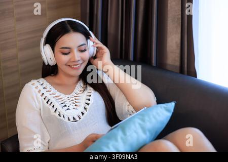 Eine glückliche junge Frau, die kabellose Kopfhörer trägt, hört Musik, während sie zu Hause auf einer Couch sitzt und ein digitales Tablet in einer Lifestyle-Umgebung hält. Stockfoto