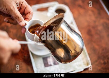 Nahaufnahme von Händen, die reichen dunklen türkischen Kaffee aus einem glänzenden Stahltopf in eine kleine weiße Keramiktasse gießen. Im verschwommenen Hintergrund ein Tablett mit anderen Stockfoto