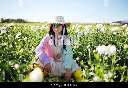 Asiatische Frau mit Sonnenbrille und Strohhut sitzt zwischen Gänseblümchen auf einem sonnigen Feld, präsentiert ein farbenfrohes Outfit und genießt das warme Wetter Stockfoto