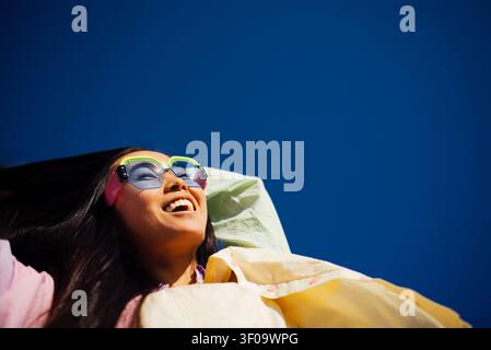 Junge Frau mit langen Haaren in bunten Sonnenbrillen liegt auf einer Decke, lächelt vor einem hellblauen Himmel, genießt die Wärme eines sonnigen Tages Stockfoto