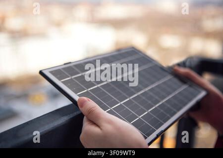 Person greift eine kompakte Solarzelle auf einem Balkon Geländer, mit einem weich ausgeblendeten Blick auf Stadtgebäude und urbane Szenerie im Hintergrund Stockfoto