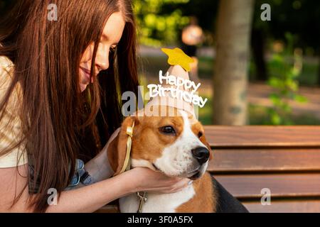 Junge Frau hält sanft ihren Beagle, der einen Happy-Birthday-Partyhut trägt, während sie gemeinsam Zeit im Freien auf einer Parkbank im warmen Abendlicht verbringen. Stockfoto
