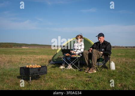 Paar arbeitet remote von einer sonnigen Wiese aus, Laptop und Telefon auf dem Schoß, grillt über einem Grill neben ihrem Zelt, genießt die Freiheit als Digital Nomads und o Stockfoto