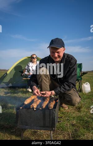 Mann, der an einem tragbaren Grill Würste über Glut und Rauch brät, während die Frau in einem Campingstuhl neben einem Zelt auf einer Wiese entspannt Stockfoto
