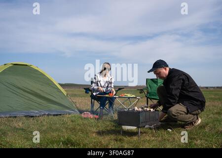 Glückliche Paare verbringen gemeinsam Freizeit auf einem Feld, grillen Essen auf einem Grill, sitzen auf Campingstühlen, genießen eine entspannende Mahlzeit im Freien unter Stockfoto