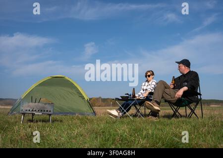 Glückliches Paar entspannt in Campingstühlen auf einer grünen Wiese, hält Bierflaschen und lächelt, während es ein Lagerfeuer für ein Picknick im Freien unter einem klaren Himmel vorbereitet Stockfoto