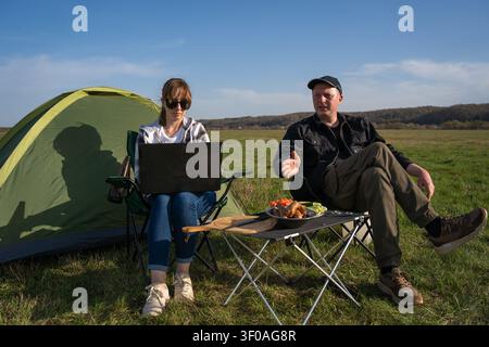 Paare campen in einer grünen Glade: Frau, die an einem Laptop an einem Picknicktisch arbeitet, während sich der Mann am Grill entspannt, sonnige Natur genießt, fernarbeitet und Stockfoto