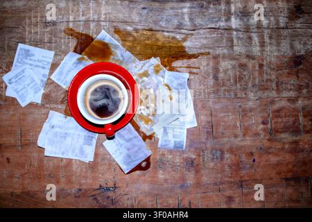 Eine rote Kaffeetasse mit schwarzem Kaffee steht auf einem Holztisch, umgeben von zerknitterten Quittungen und einem Kaffeefleck, der eine unordentliche Szene erzeugt Stockfoto