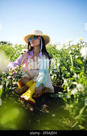 Asiatische Frau in lebhafter Kleidung und gelben Stiefeln posiert in einem Blumenfeld, trägt einen Strohhut und eine Sonnenbrille vor einem strahlend blauen Himmel Stockfoto