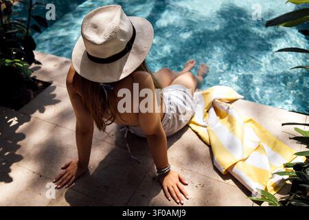 Frau mit Sonnenhut sitzt am Schwimmbecken, trägt helle Kleidung, mit einem Handtuch neben ihr und üppiger Vegetation im Hintergrund Stockfoto