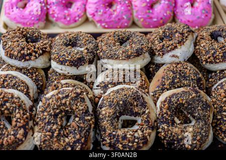 Frische schokoladige erdnussglänzende Donuts gestapelt zur Schau in einer thailändischen Bäckerei mit farbenfrohen Gebäck im Hintergrund und flacher Schärfentiefe. Stockfoto