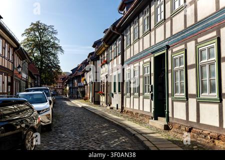 Sonnenbeschienene Wernigeröder Wahrzeichenstraße windet sich durch Holzhäuser, Kopfsteinpflaster, geparkte Autos und blumige Fassaden. Historische Harz-Stadtszene zeigt Deutsch Stockfoto