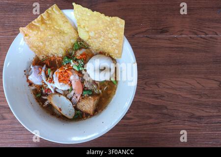 Oberansicht von authentischen thailändischen Tom-Yum-Meeresfrüchte-Nudeln mit Garnelen, Tintenfisch, Fischbällchen, Kräutern, Erdnüssen und Kopierraum auf Holztablett. Stockfoto