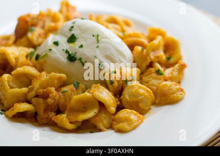 Nahaufnahme von Orecchiette in Orangensauce, garniert mit einer ganzen Burrata und gehackter Petersilie, serviert auf einem weißen Teller, Bambusmattenrand, apulische Küche Stockfoto