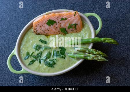 Oberansicht einer cremigen Erbsensuppe mit Lachsfilet, Dill, Petersilie und drei Spargelstangen in einer ovalen Schale mit grünen Griffen auf dunklem Granit Stockfoto