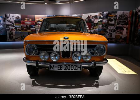 München, Deutschland – 7. Juli 2026: Vorderansicht eines orangefarbenen klassischen BMW 2002ti, der in einem Museum ausgestellt ist, zeigt Kühlergrill, Scheinwerfer und historische Fotokollage Stockfoto