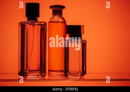 Drei Glasparfümflaschen mit schwarzen Verschlüssen sind auf einem Regal angeordnet, vor einem warmen orangefarbenen Hintergrund, was ihre einzigartigen Formen und Größen betont Stockfoto