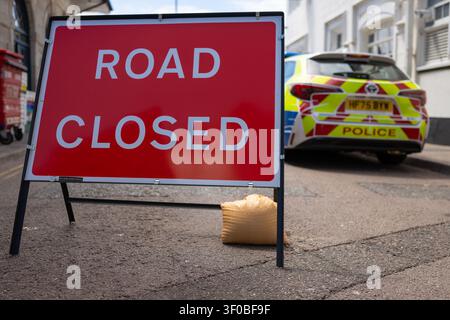 Ein rotes Straßensperrungsschild auf einer Straße mit einem verschwommenen britischen Polizeiauto, das die Straße während einer Polizeisperre oder eines Verkehrsunfalls blockiert, Vereinigtes Königreich. Stockfoto