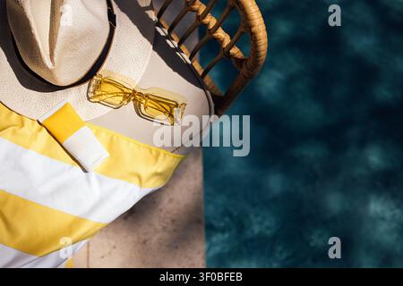 Pool-Side-Einrichtung mit einem Strohhut, gelber Sonnenbrille und Sonnencreme auf einem gestreiften Handtuch neben einem klaren blauen Schwimmbecken Stockfoto