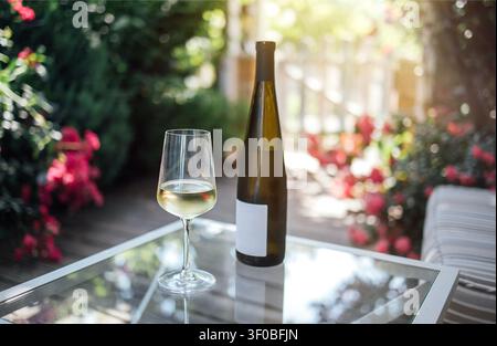 Glas weißer Wein neben einer Flasche auf einem Glastisch in einem Garten voller lebendiger Blumen und üppiger Grünpflanzen im Sonnenlicht Stockfoto