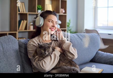 Glückliche junge Frau mit Kopfhörern, die eine Katze auf dem Sofa hält und zu Hause entspannt Stockfoto