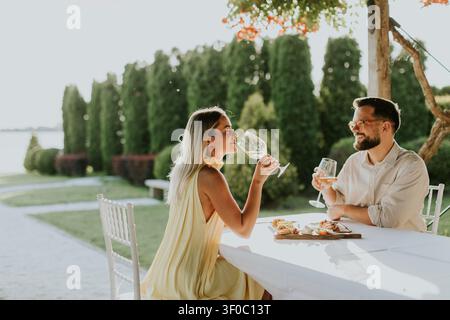 Zwei Personen sitzen an einem Tisch in der Nähe eines Sees. Sie lächeln, während sie aus Gläsern trinken und Essen teilen. Die Sonne scheint auf ihre Gesichter, während sie es genießen Stockfoto
