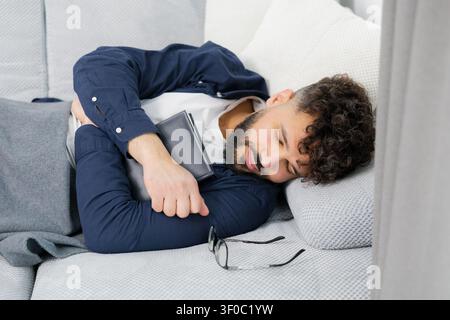 Junger Mann schläft friedlich auf einer bequemen Couch, macht einen Tagesausruhen nach dem Lesen, fühlt sich entspannt und ruhig Stockfoto