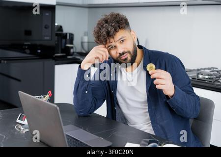 Mann mit lockigen Haaren hält eine Bitcoin-Münze, sitzt an einem Tisch mit einem Laptop und verwaltet Kryptowährungsinvestitionen von zu Hause aus Stockfoto