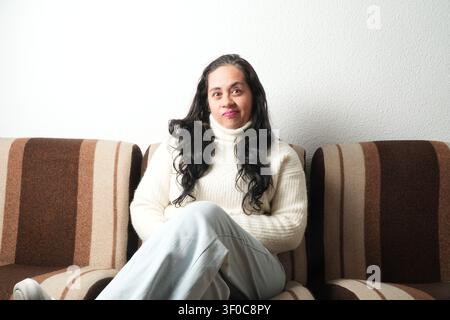 Frau sitzt bequem auf einem gestreiften Sofa. Sie trägt einen weichen, cremefarbenen Rollkragenpullover. Langes, dunkles Haar fließt locker um ihre Schultern. Sie lächelt g Stockfoto
