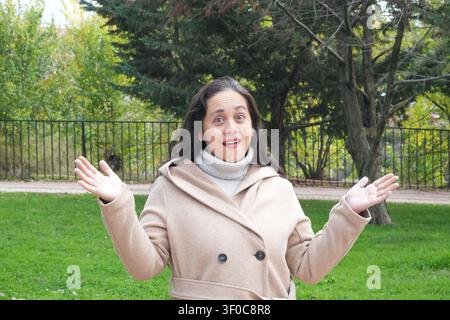 Frau lächelt im Park, Arme weit geöffnet. Sie trägt einen beigen Mantel und einen grauen Schal. Bäume und grünes Gras umgeben sie. Parkzaun im Hintergrund sichtbar. Hellig Stockfoto