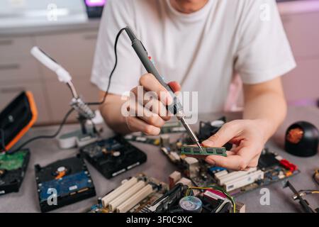 Nahaufnahme von Technikerhänden, die mit einem Lötkolben ein grünes Computer-RAM-Modul reparieren. Werkbank übersät mit Mainboards und anderen elektronischen Bauteilen Stockfoto