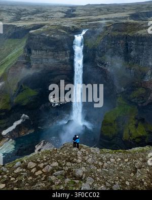 Aus der Vogelperspektive des Haifoss-Wasserfalls, der über moosige Klippen stürzt, mit einem Paar, das am felsigen Rand in der südlichen Region, Island, steht. Stockfoto