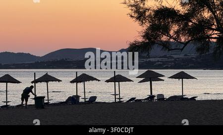 Abendliche Strandszene mit einer Person, die am Ufer aufräumt, silhouettiert vor einem ruhigen Sonnenuntergang mit Sonnenschirmen und Liegestühlen am Wasser, schaffend Stockfoto