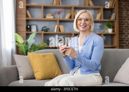 Zufriedene reife Frau hält ein Smartphone und lächelt in die Kamera, während sie sich auf einer bequemen Couch in ihrem modernen Wohnzimmer entspannt Stockfoto