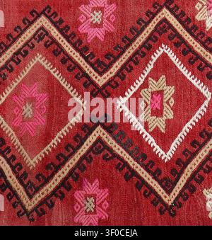 Roter Kilim-Teppich, der eine reiche Stickerei traditioneller geometrischer Muster zeigt, mit markanten Diamantformen und stilisierten rosa Blumenelementen erneut Stockfoto