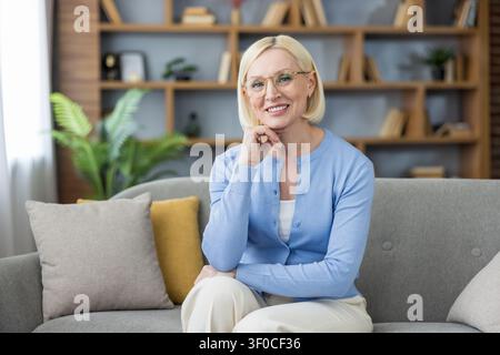 Reife blonde Frau mit Brille und blauem Cardigan, die bequem auf einem modernen Sofa sitzt und in einem entspannten Wohnumfeld in die Kamera lächelt Stockfoto