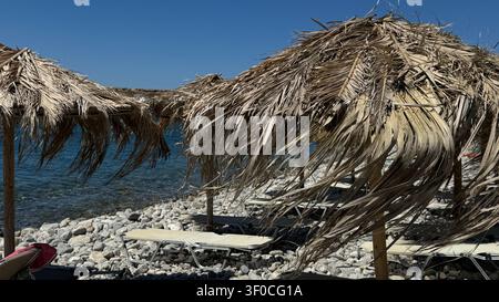 Strohschirme und Liegestühle an einem felsigen Strand bieten Meereserholung bei klarem Himmel und ruhiger Meereskulisse. Ideale Szenerie für einen friedlichen Strand Stockfoto