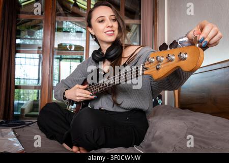 Junge Musikerin stimmt eine E-Bassgitarre, während sie Kopfhörer in einem Heimstudio trägt Stockfoto
