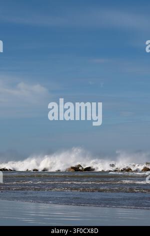 Kraftvolle Wellen schlagen gegen die felsige Küste. Ein klarer blauer Himmel erstreckt sich darüber mit dünnen Wolken. Wasser sprüht in die Luft, während die Flut hereinrollt. Ozean mich Stockfoto