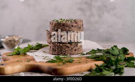 Ein vertikaler Stapel roher schwarzer Bohnenburger-Patties ist mit Samen und frischer Petersilie auf Pergamentpapier garniert. Das rustikale Holzbrett und das weiche, diffuse Stockfoto