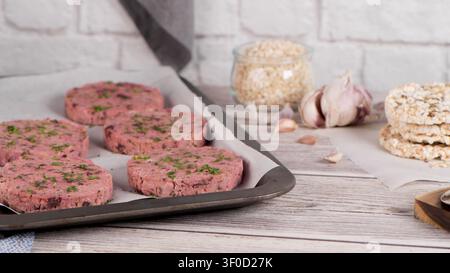 Mehrere rohe pflanzliche Burger-Patties, die mit frischen grünen Kräutern belegt sind, ruhen auf Pergamentpapier. Die rustikale Szene spielt sich auf einer hellen Holzoberfläche mit Garne ab Stockfoto