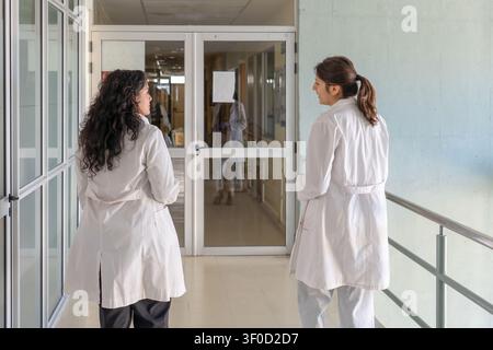 Rückansicht von zwei jungen Frauen in weißen Laborkitteln, die in einem hellen institutionellen Korridor stehen und sich in der Nähe von Glastüren unterhalten, mit einer weiteren Person Stockfoto