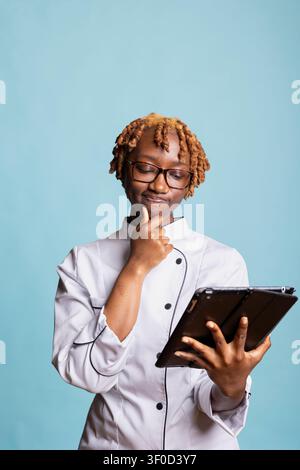 Nachdenklicher Koch mit Hand am Kinn liest Online-Rezept, plant Anweisungen und recherchiert Ideen für Kochbuch. Professioneller Koch überprüft Details auf Tablet, bereitet sich darauf vor, Gourmet-Mahlzeit zuzubereiten. Stockfoto