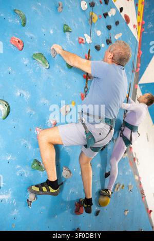 Älterer Mann im Gurtzeug mit Sicherheitsseil, der an einer Boulderwand klettert Stockfoto